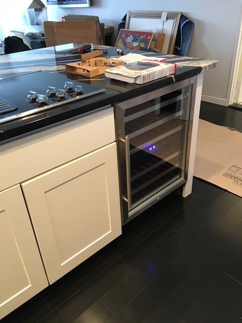 Wine cooler built into a white cabinet, installed in a kitchen with dark flooring.