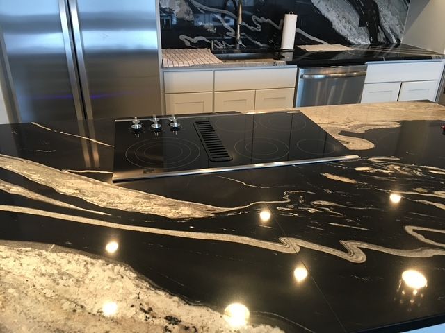 Black granite kitchen island with built-in cooktop, white streaks, stainless steel appliances, and white cabinets.