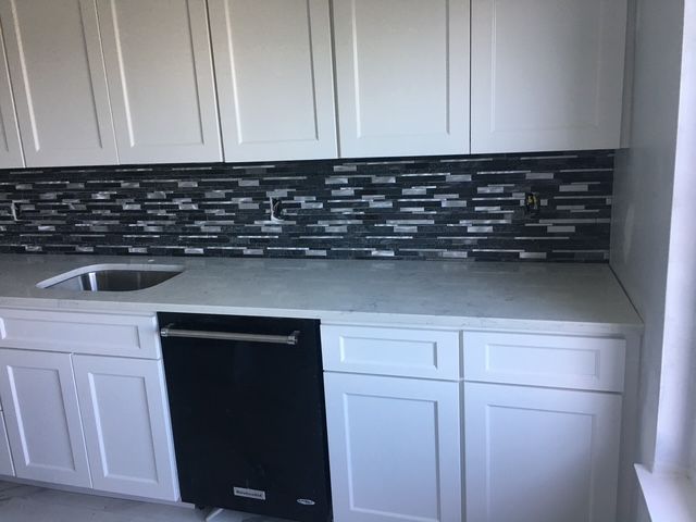 White kitchen cabinets with a dark tiled backsplash, gray countertop, and black dishwasher.