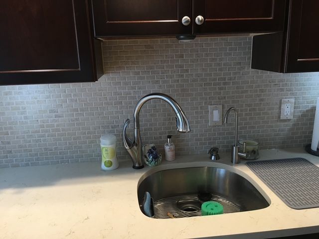 Kitchen sink with a chrome faucet and white countertop, beige tile backsplash, and dark brown cabinets.