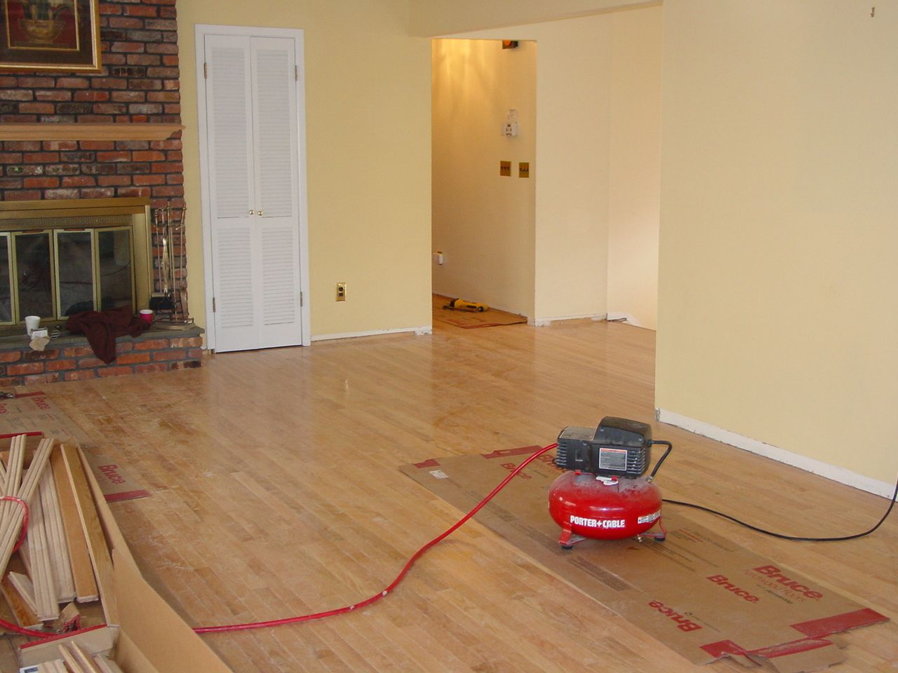 Hardwood floor being installed in a living room, red air compressor in center, near a brick fireplace.