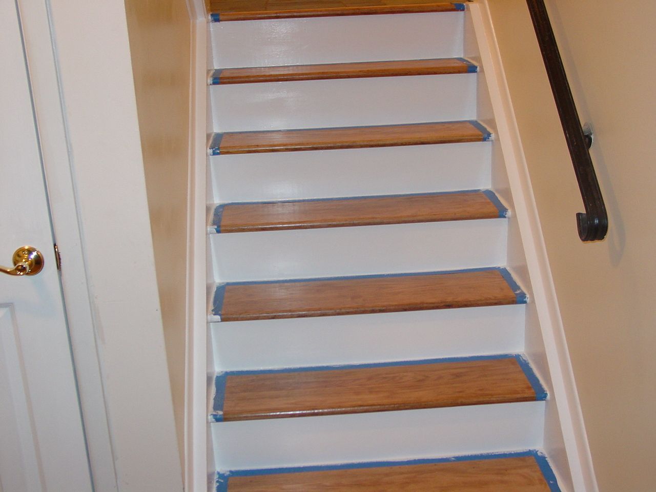 Staircase with wooden treads and white risers, blue tape along edges, black handrail on right.