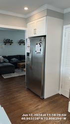 Stainless steel refrigerator with white cabinets, wood floor, and view into living room.