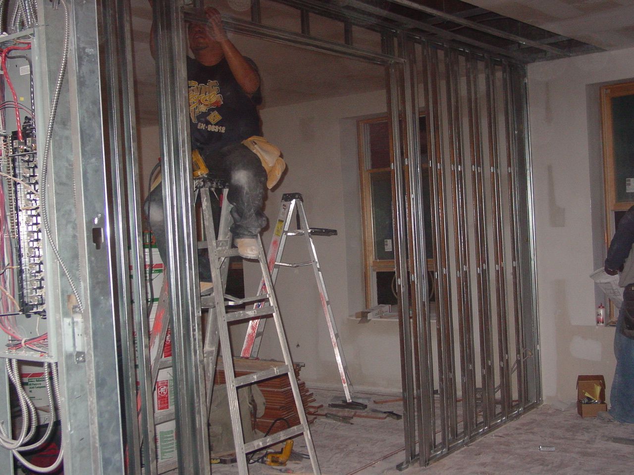 Construction worker on a ladder framing a metal interior wall. Electrical panel on the left, window in the background.