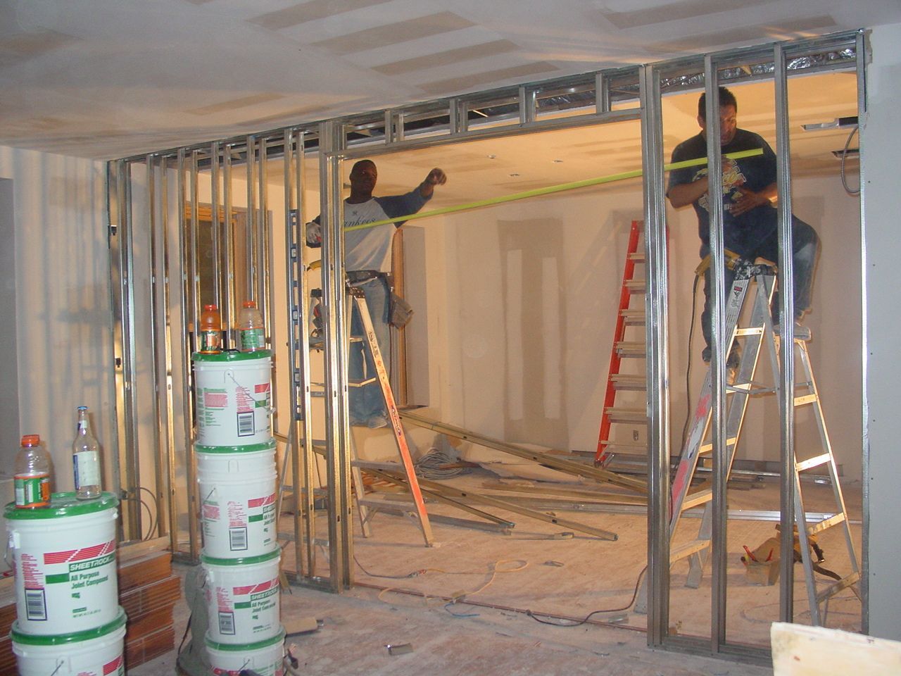 Construction workers framing a doorway in a room. Metal studs and drywall visible.