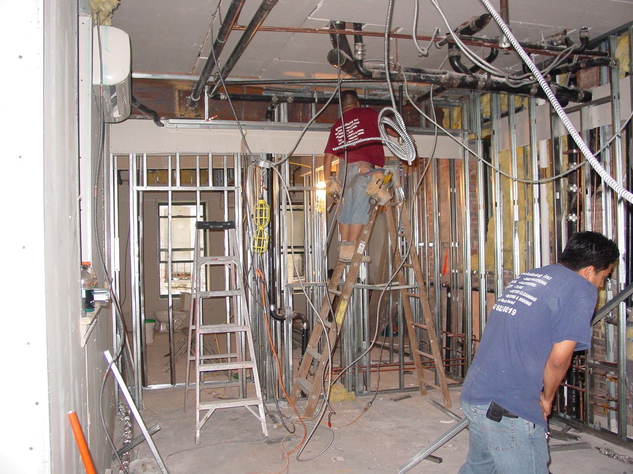 Construction workers in a room with metal framing and exposed wiring, one on a ladder, one working on the wall.