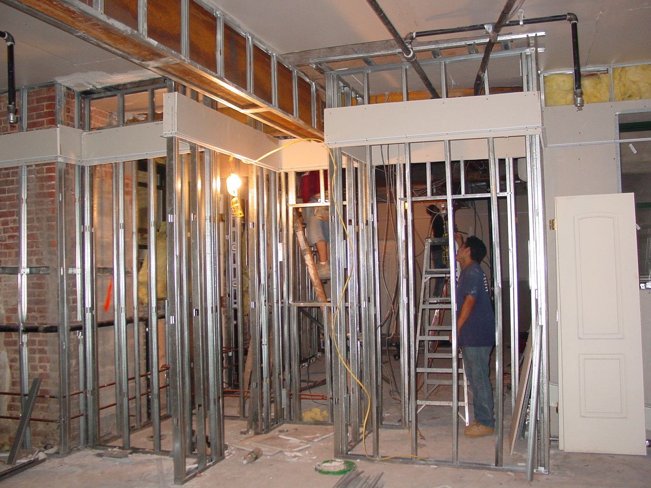 Construction site: metal stud framing for walls, two workers, brick wall visible, exposed pipes.