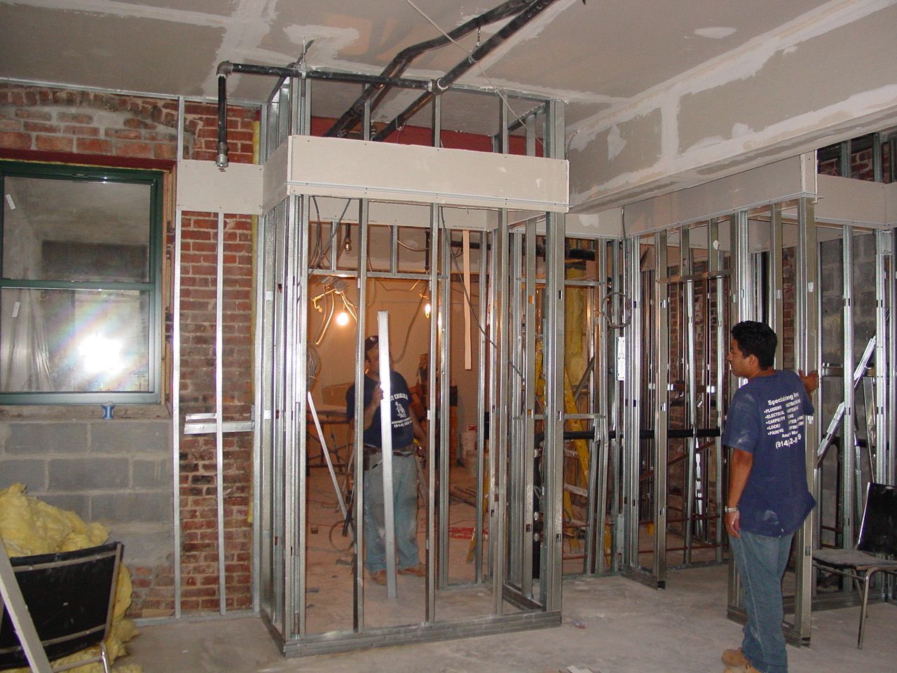 Construction site: metal framing for doorways, workers, exposed brick wall, wiring, and drywall.