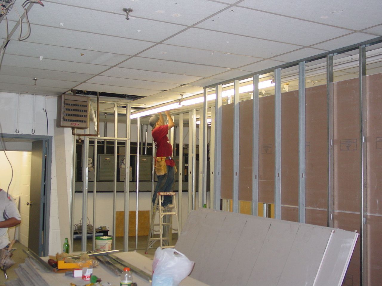 Construction worker on ladder installing drywall in a room with metal framing.