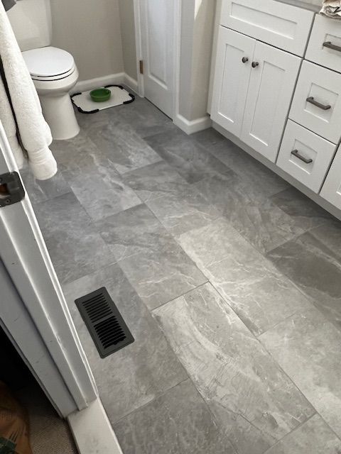 Gray marble-look tiled bathroom floor with white vanity and toilet. Black vent on the floor.
