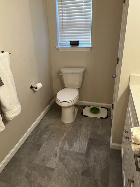 Small bathroom with a toilet, window, and grey tile flooring. Beige walls and white trim.
