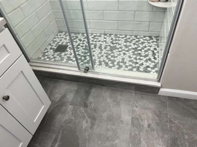 Bathroom with gray marble-look floor, pebble shower floor, glass shower door, white cabinet, and light gray walls.