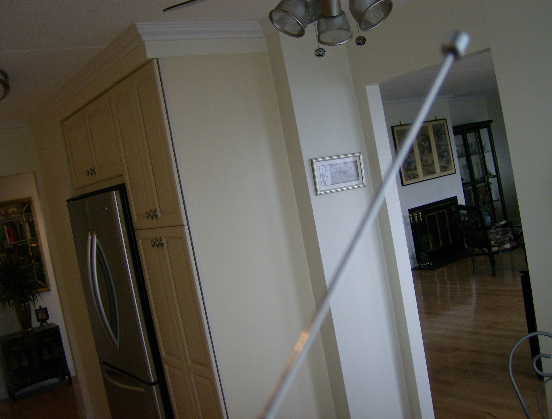 Kitchen interior with tall, metallic rod in the foreground; refrigerator, cabinets, and doorway visible.