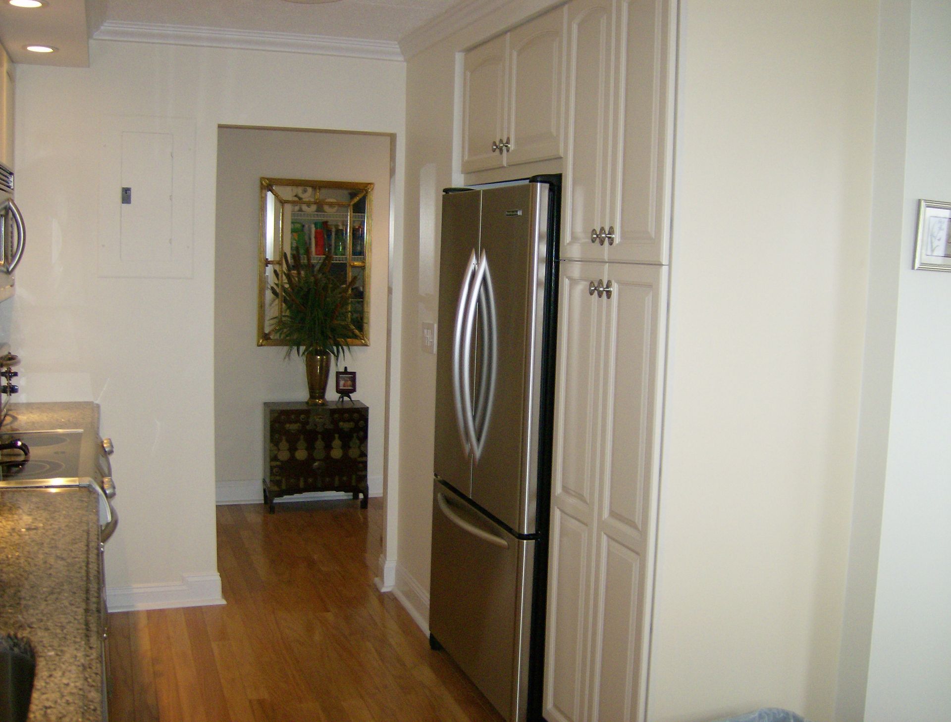 Kitchen with stainless steel refrigerator, beige cabinets, and view into a hallway with wooden floor.