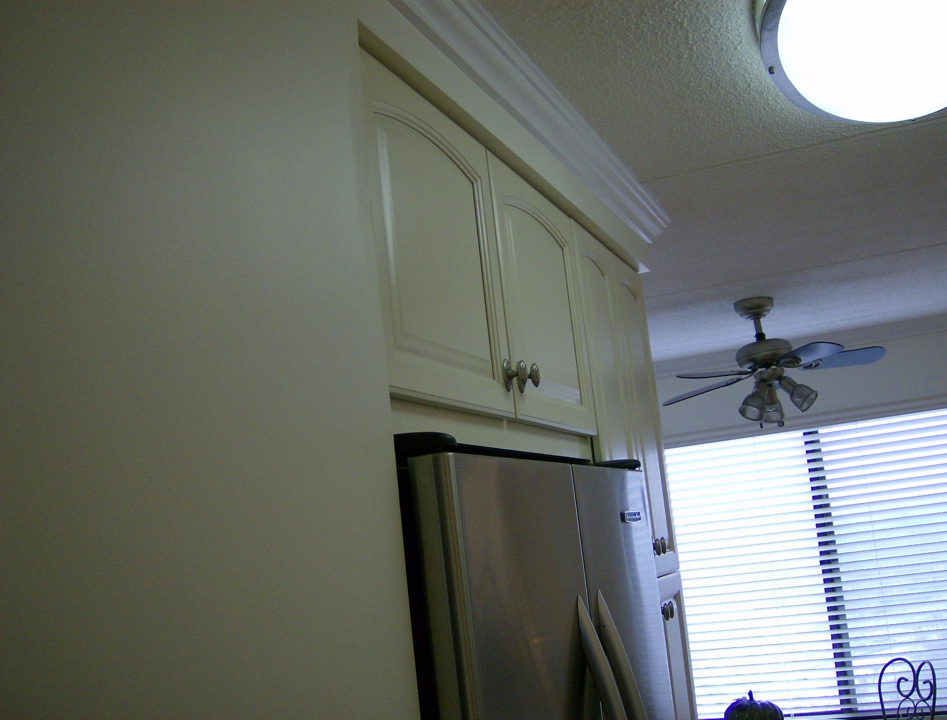 White kitchen cabinets with crown molding above a stainless steel refrigerator, near a window and ceiling fan.