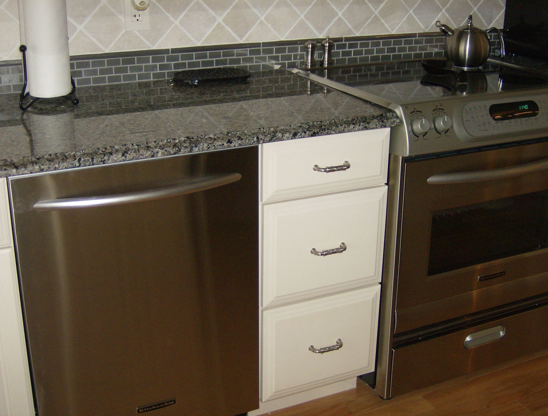 Stainless steel dishwasher and oven flanking white cabinet with drawers under granite countertop in kitchen.