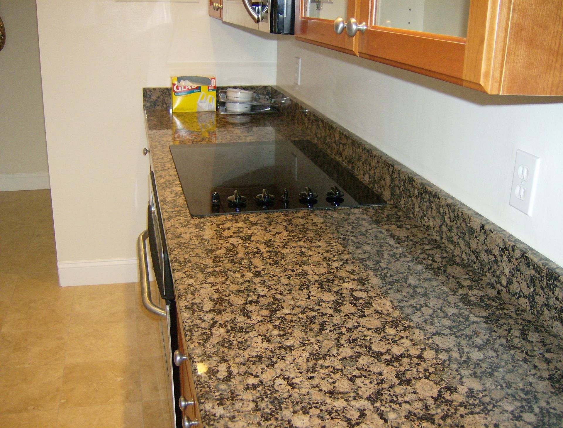 Granite countertop with built-in black stovetop, beneath wooden cabinets, near an outlet and beige wall.