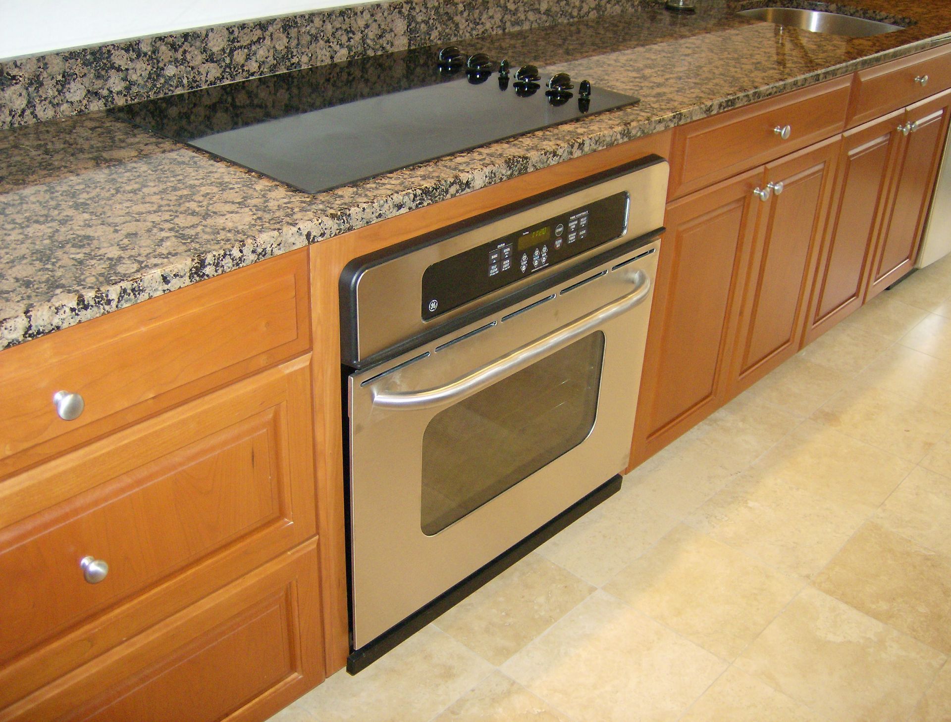 Stainless steel oven built into wooden cabinets under a speckled countertop with stovetop.