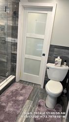 A bathroom with a frosted glass door, white toilet, purple rug, and gray tiled walls.
