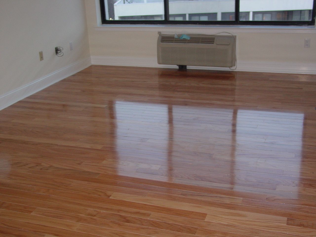 Shiny hardwood floor reflects a window. Beige walls, white trim, and a window AC unit are present.