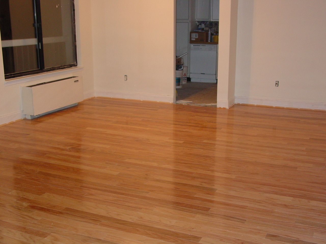 Empty room with shiny wood floors, white walls, and an open doorway leading to a kitchen.