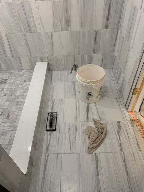 Bathroom with gray and white tiled walls. A bucket, trowel, and cloth rest on the floor.