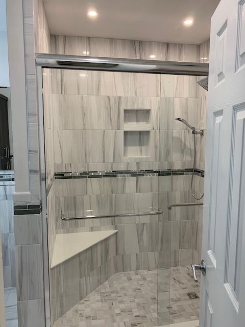 A tiled shower with glass doors and a built-in seat, featuring gray and white tones, a shelf, and a grab bar.