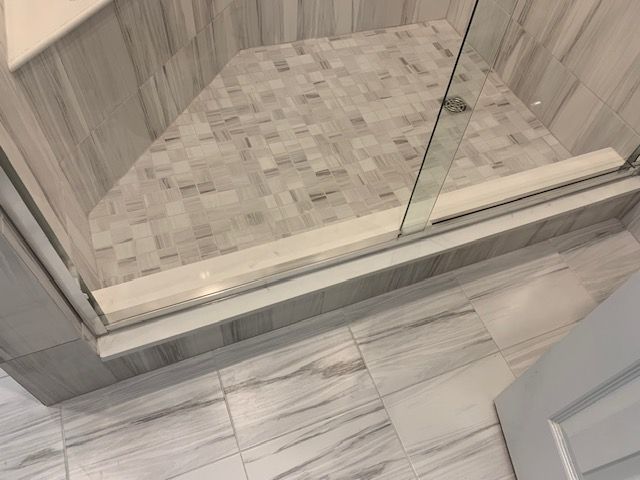 Shower with marble-look tiles. Glass door sits atop a step, tiles on floor and walls are gray/white.