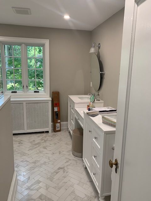 Bathroom with white cabinets, marble floors, window, and a large oval mirror.