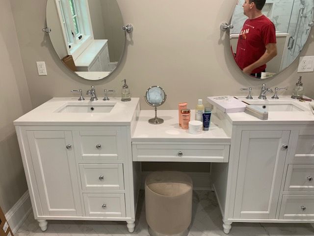 A bathroom with two white vanities, oval mirrors, and a makeup vanity. A man in a red shirt is reflected in the mirror.