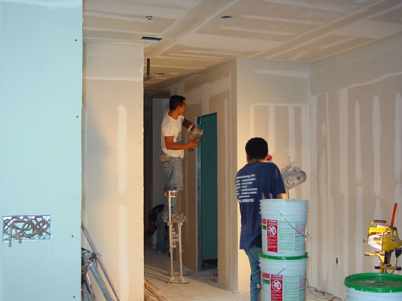 Two workers drywalling a room. One stands on stilts, the other next to paint buckets.