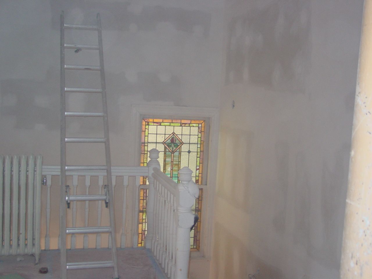 A tall aluminum ladder stands near a staircase with a stained glass window. Walls are being renovated.