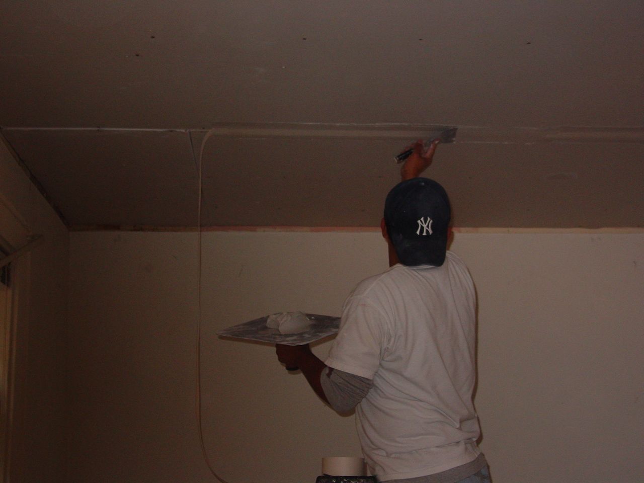 Person on a ladder, smoothing ceiling with plaster. White walls, baseball cap, plastering tools.