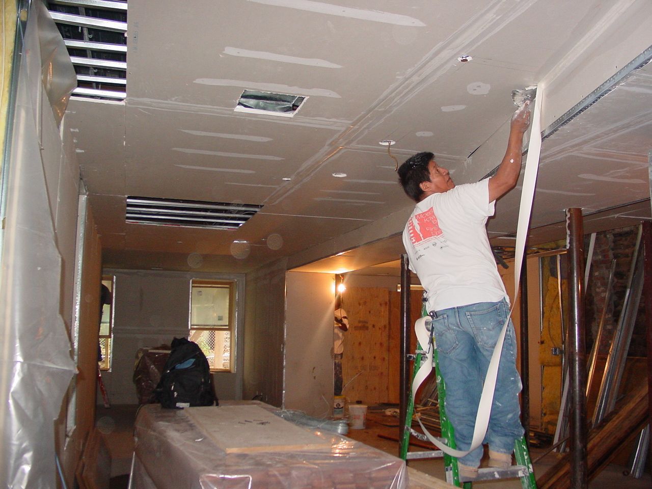 Man on ladder taping drywall ceiling in a room under renovation.