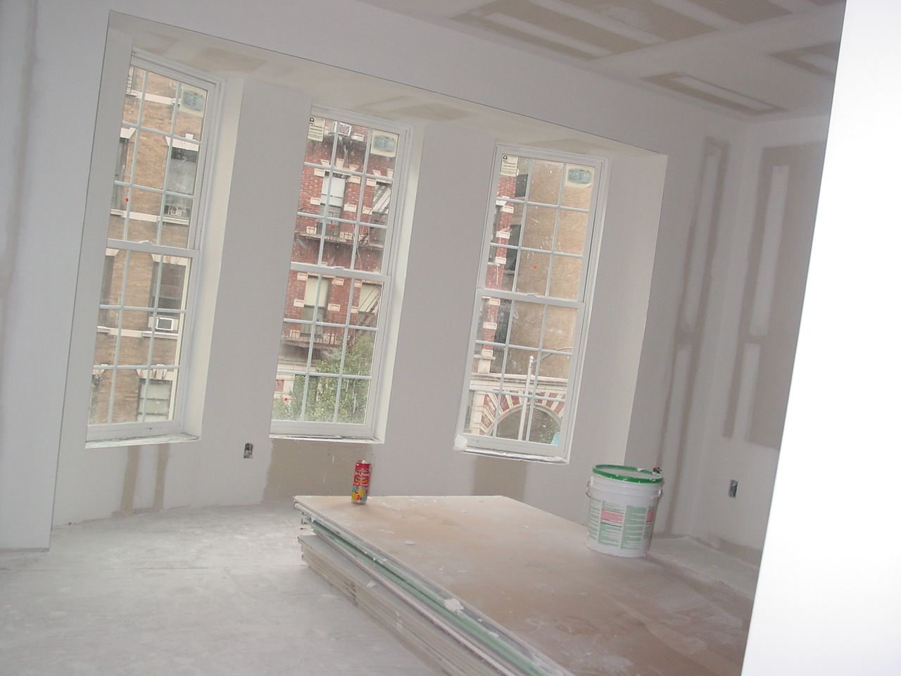 Interior room under construction with three windows overlooking a city, drywall, and supplies.
