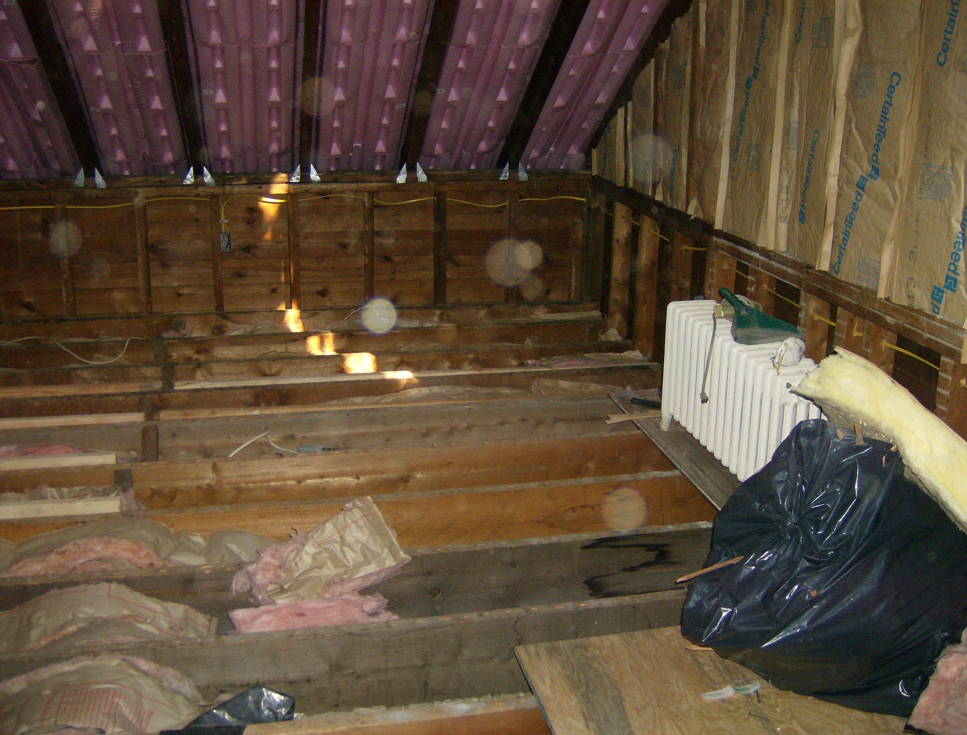 Attic space with exposed wood beams, insulation, and a radiator. A black trash bag sits near the radiator.