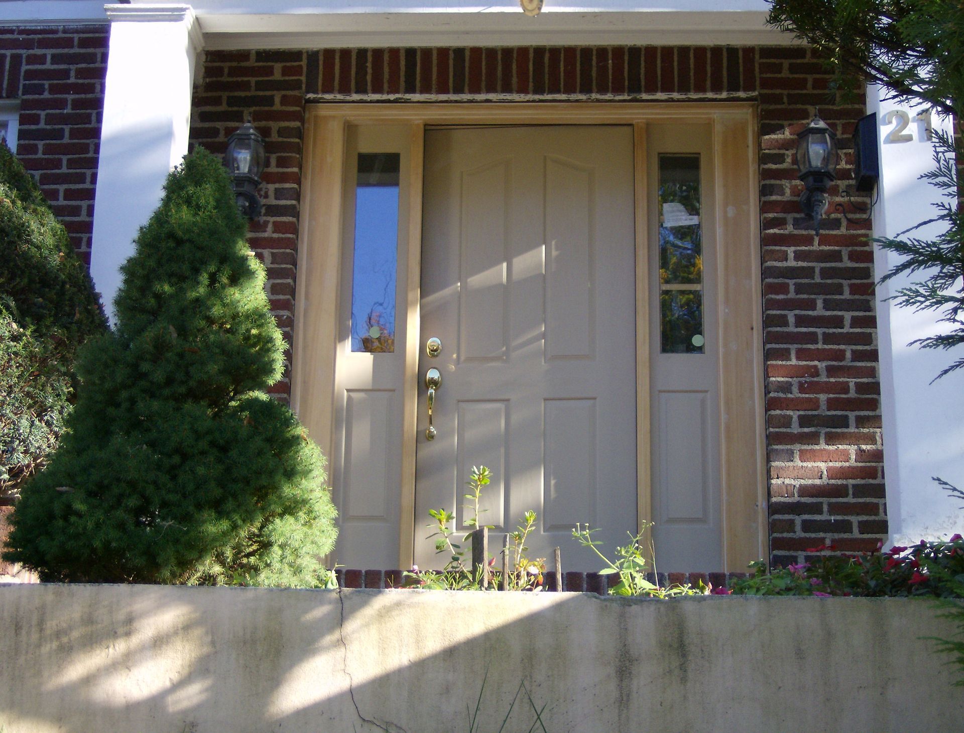 Tan front door with sidelights, flanked by brick. A small tree is on the left.