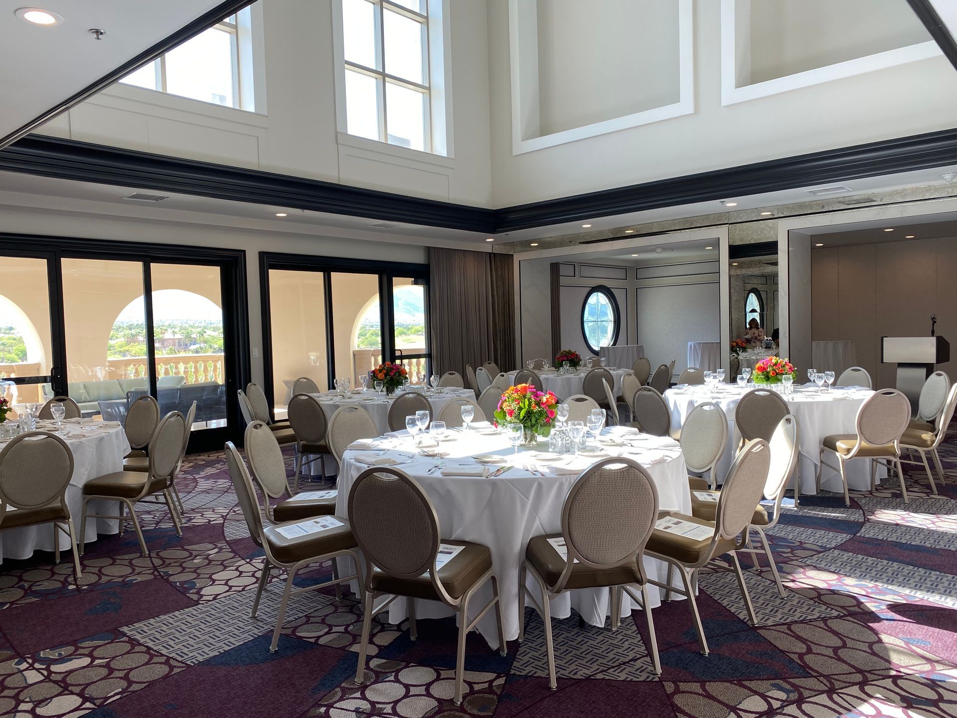 A ballroom with round tables set for a catered event, with flowers in the center and beige chairs. Large windows and ornate ceiling.
