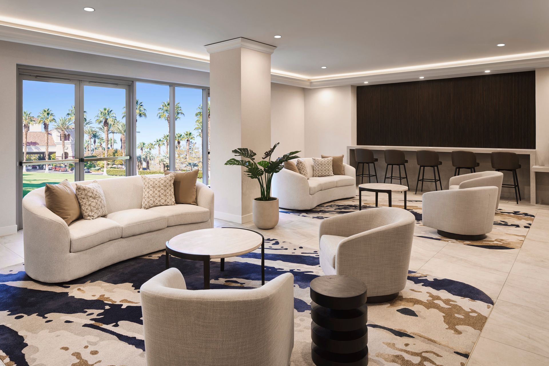 Modern lounge area with curved white sofas, armchairs, and a bar, featuring a patterned rug and large windows with palm trees.