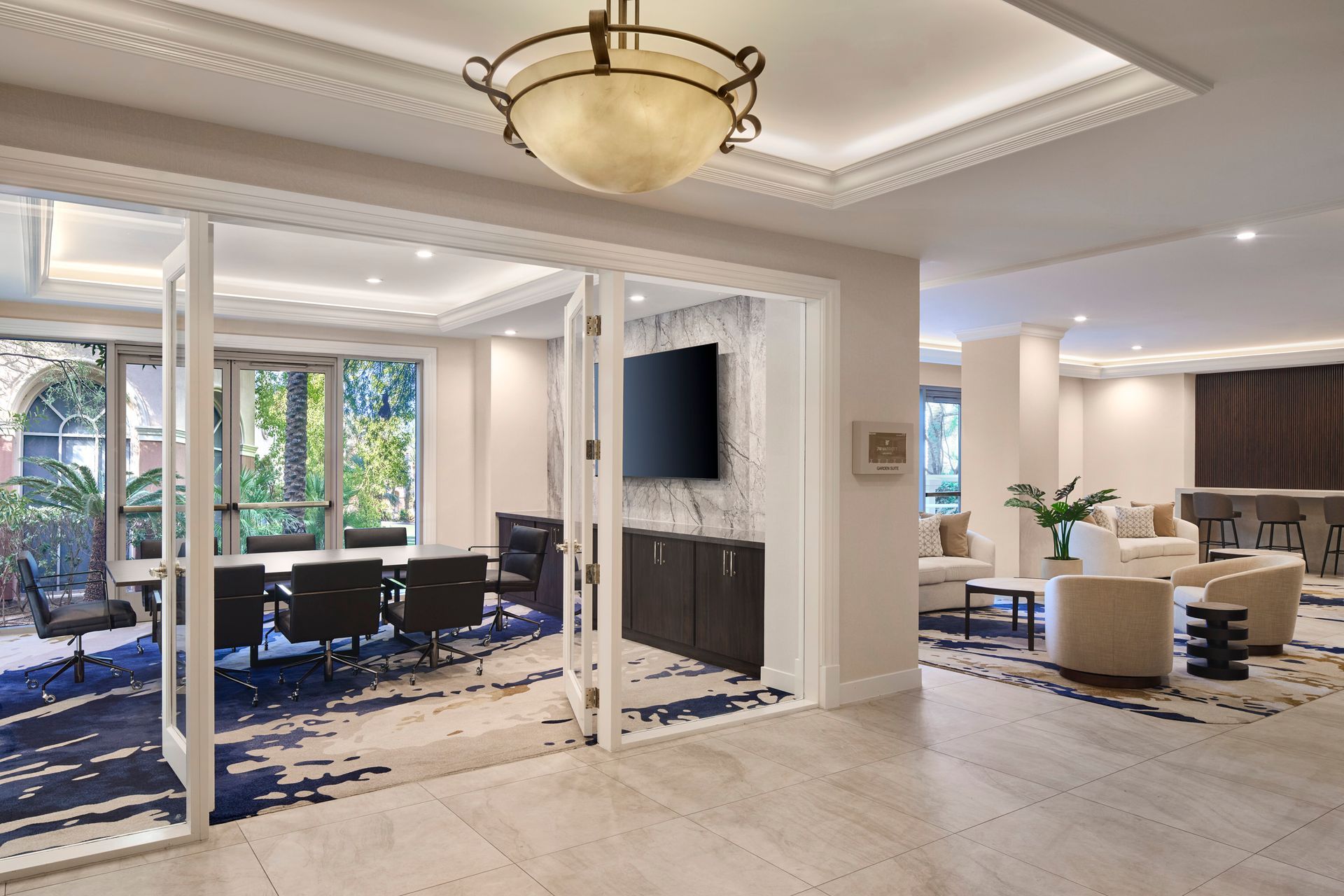 A modern hotel lobby with a glass-walled meeting room and seating area. The carpet is blue and white, the walls are light, and there is an outdoor patio visible.