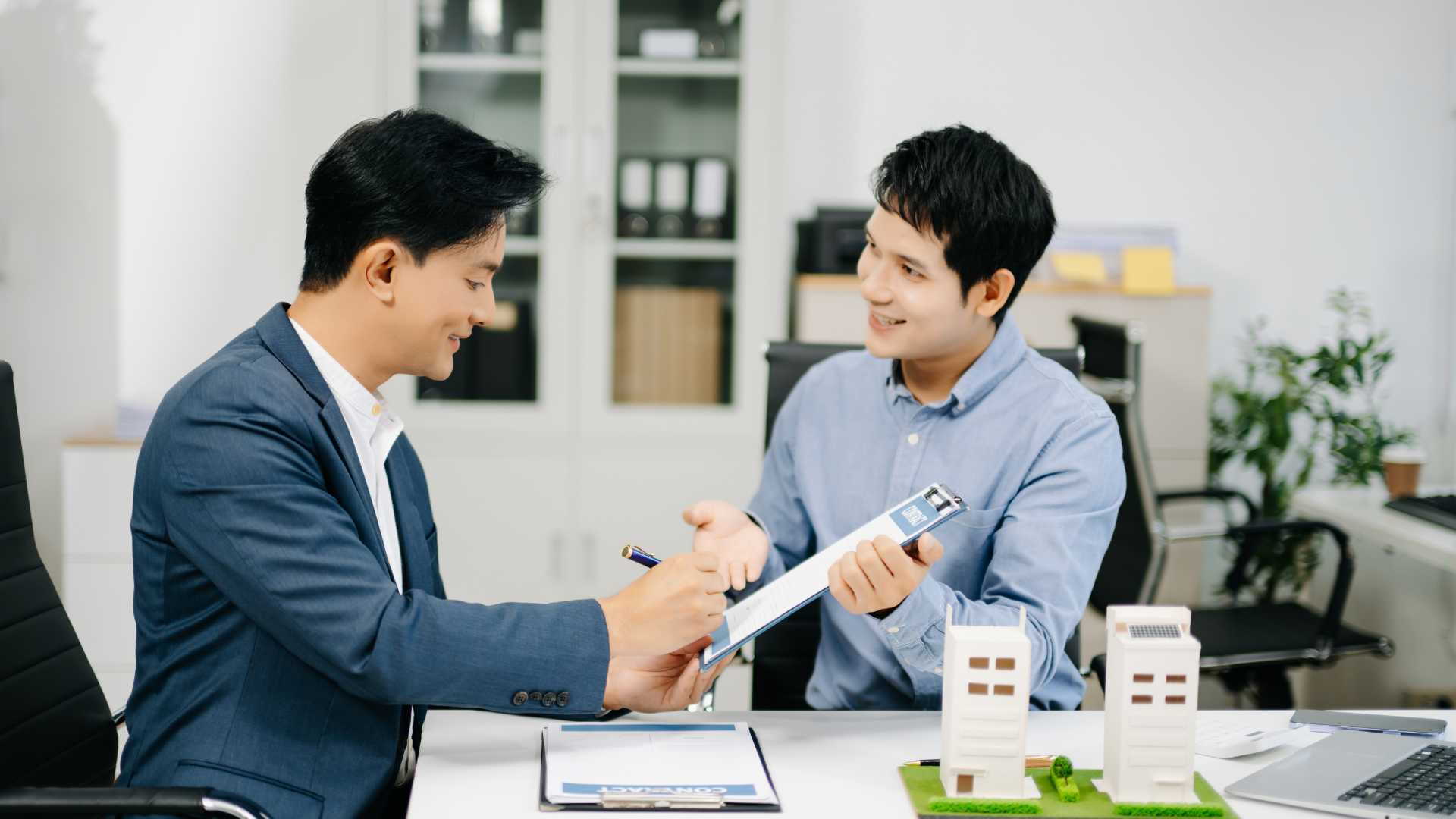 Two men discussing documents across a desk in a bright office with building models in the foreground