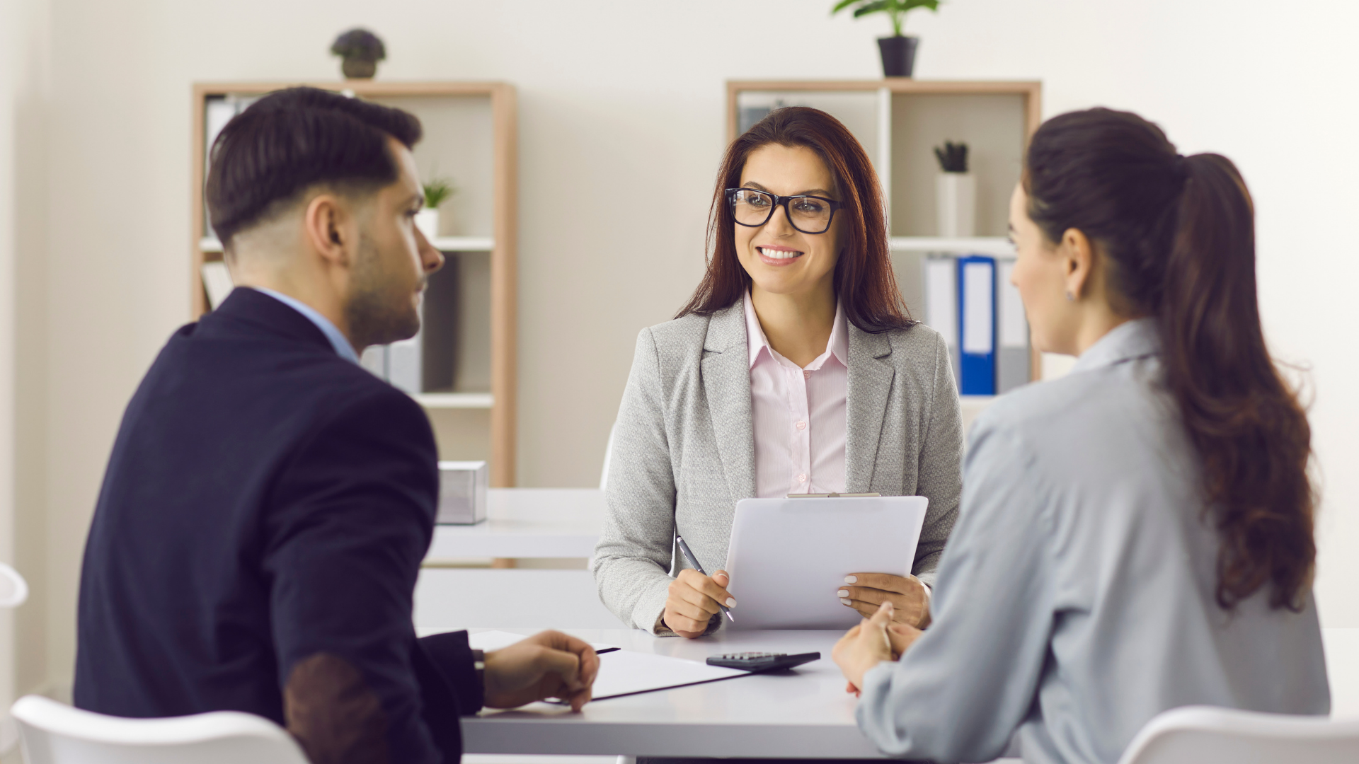 A woman in glasses smiles while holding a document and speaking to a couple at a table in an office.