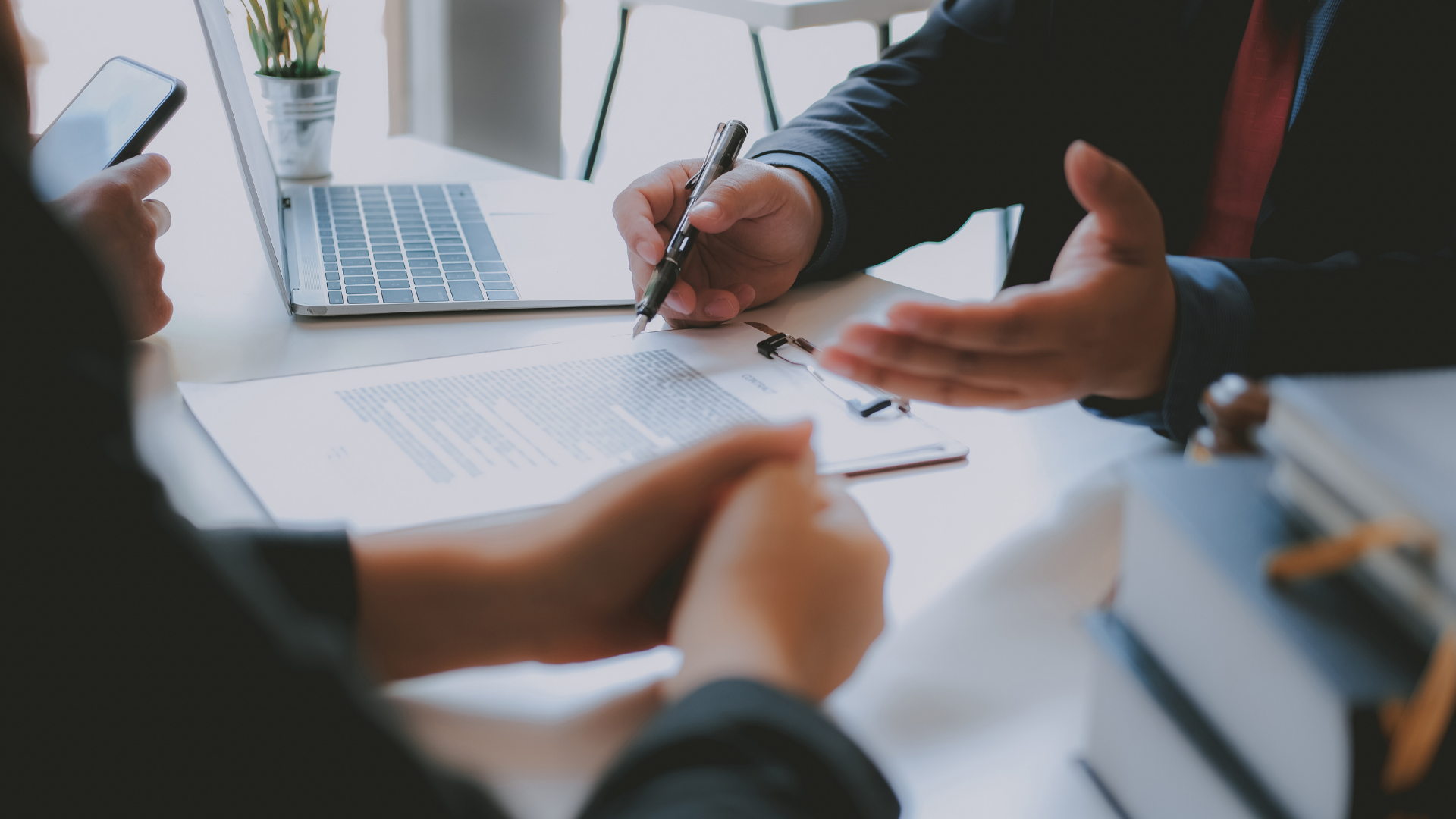 Person signing document at a table with a laptop, another person, and law books in an office setting.