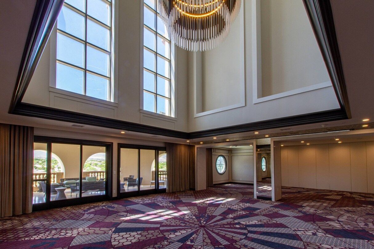 Empty event room with high ceilings, large windows, and patterned carpet. Sunlight streams in; a chandelier hangs from the ceiling.