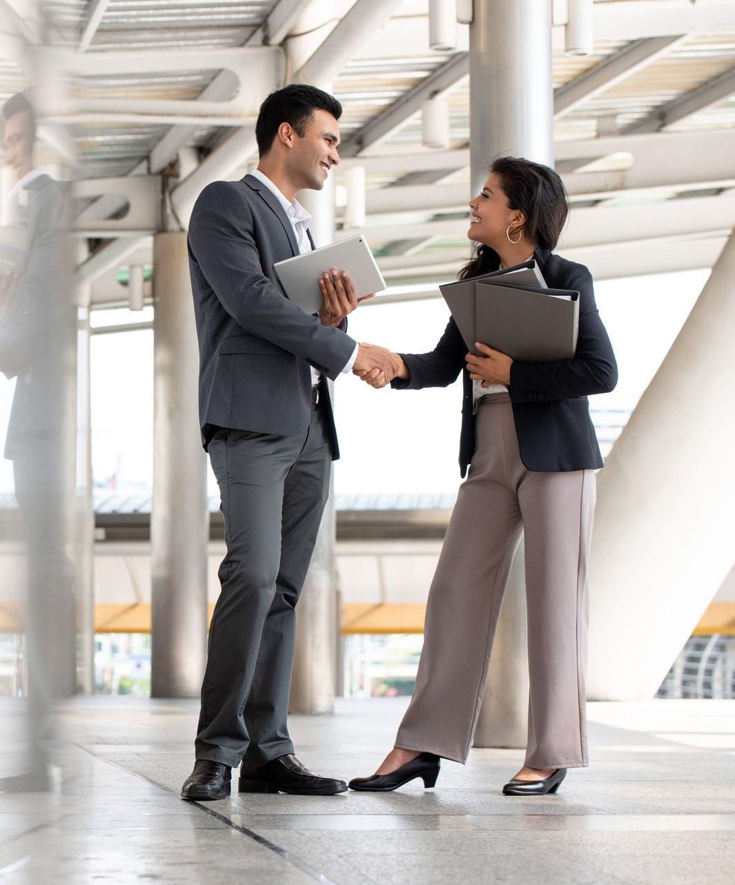 Two professionals shaking hands; a man in a suit and a woman in a blazer. They're smiling, in an outdoor urban setting.