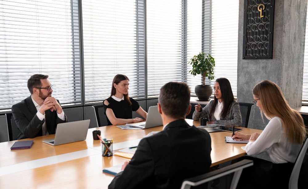 Business meeting: five people seated around a table with laptops, discussing in an office setting.