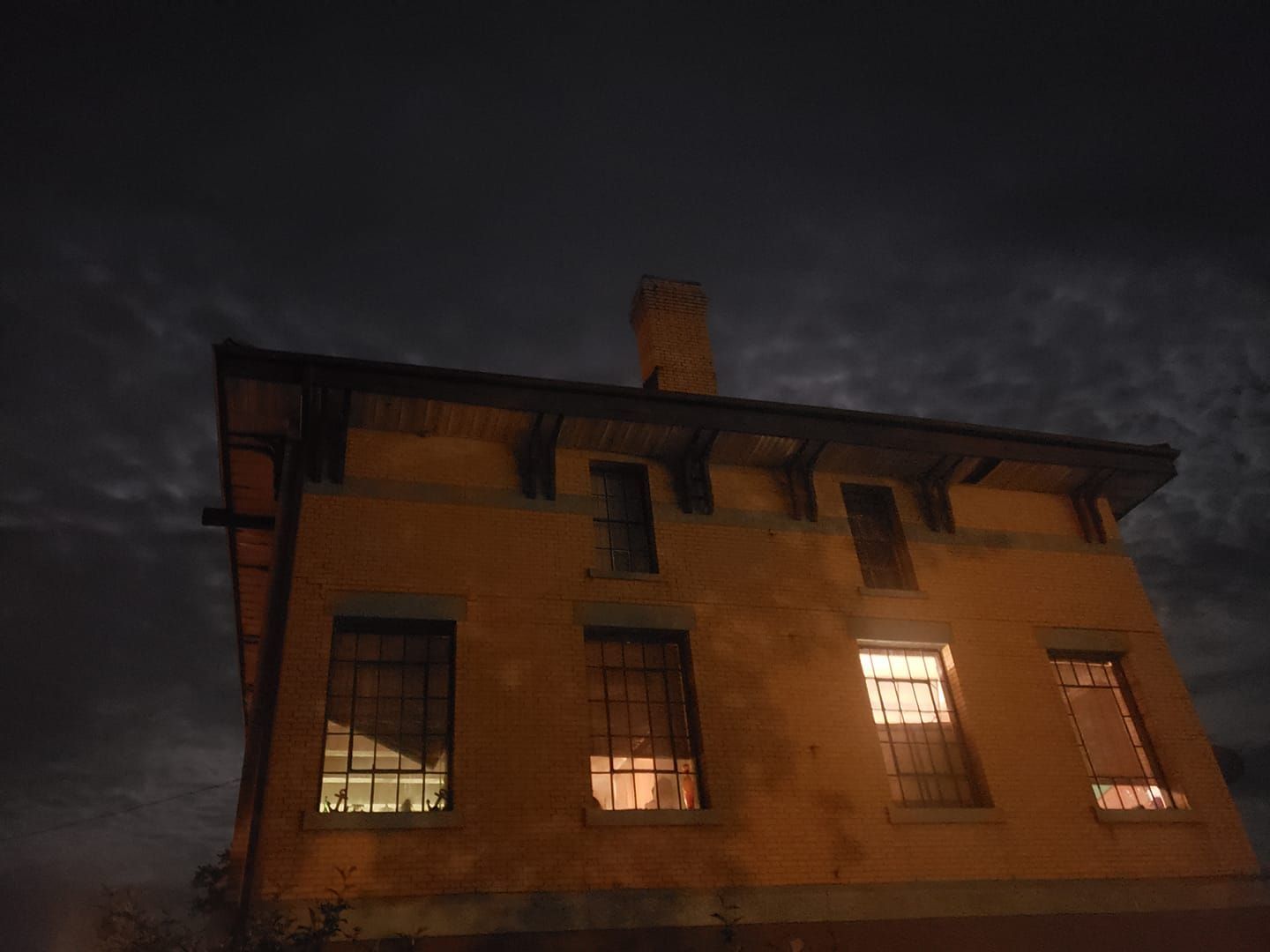 A low-angle view of a brick building at night with warm, glowing light spilling from its windows under a cloudy sky.