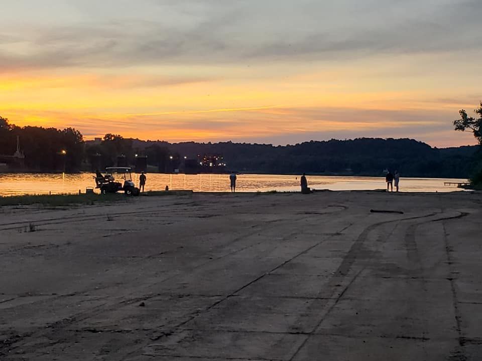 A boat ramp at sunset with silhouettes of people near the water and a vehicle parked on the concrete bank.
