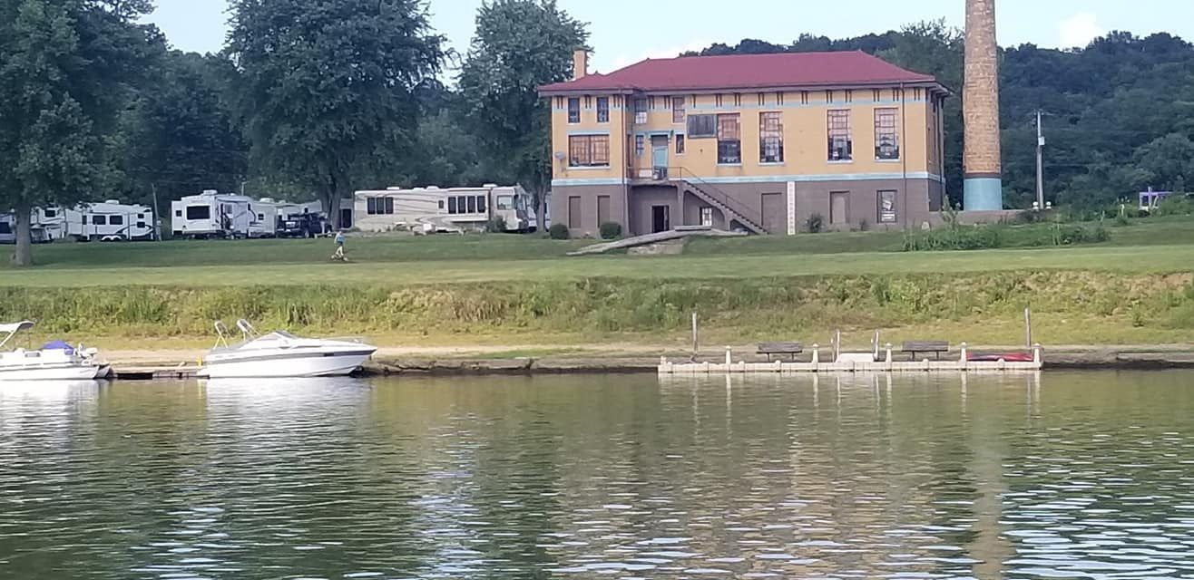 A yellow two-story building with a red roof and brick chimney sits on a grassy bank by a river with boats docked nearby.