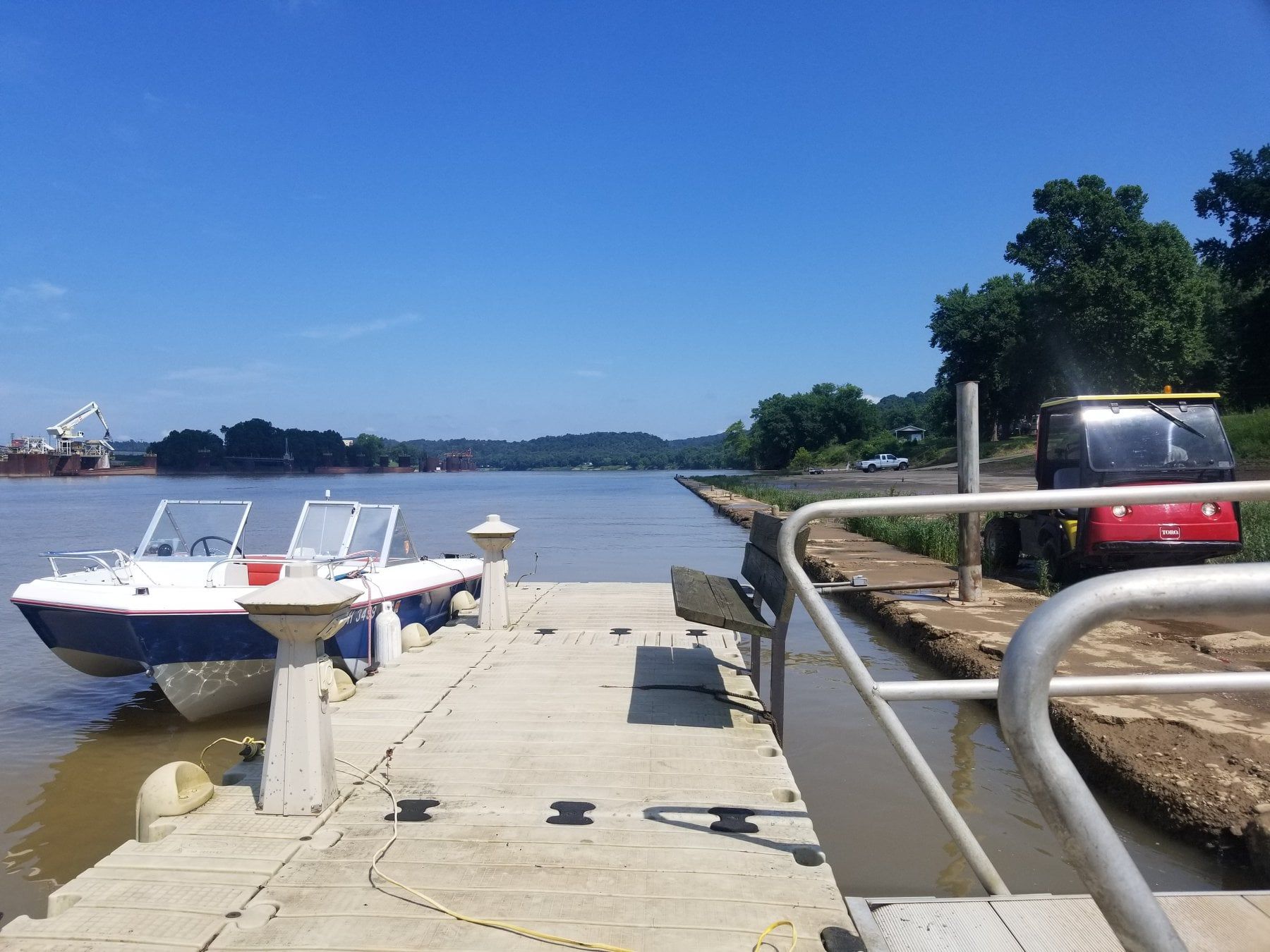 A boat moored at a concrete dock on a river under a clear blue sky, with a small utility vehicle parked nearby on land.